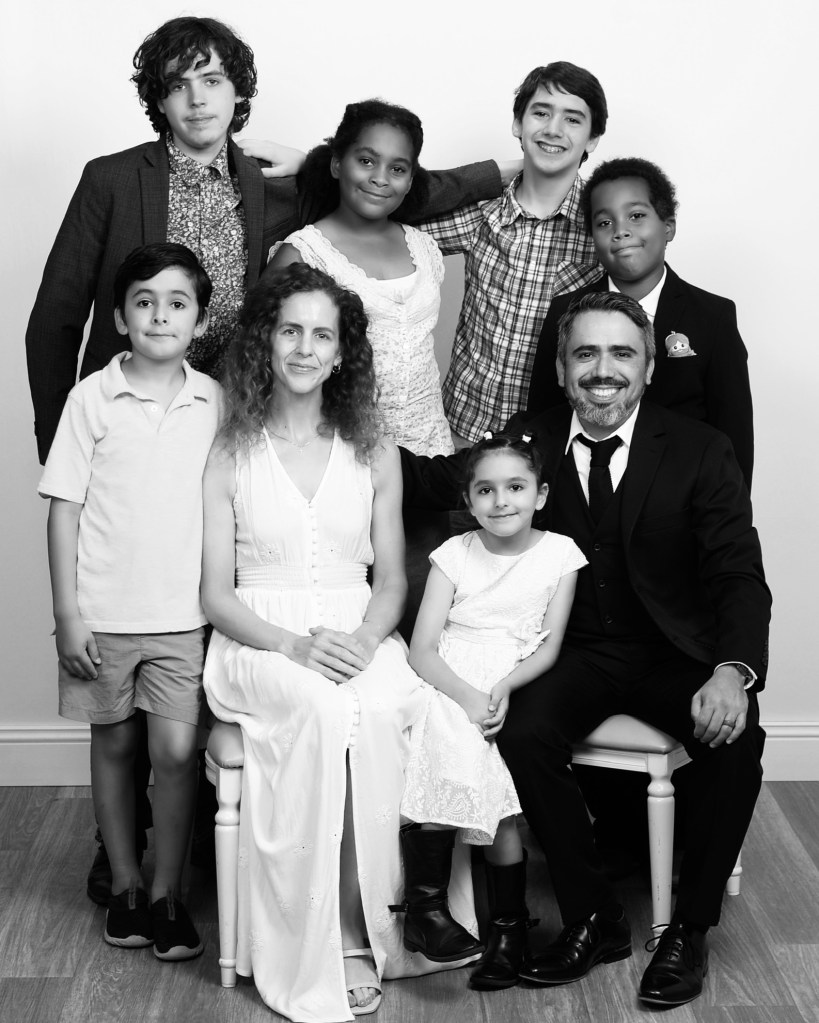 A black and white family portrait featuring two adults seated in front and six children arranged behind them. The adults smile warmly, while the children display a mix of joyful and playful expressions.
